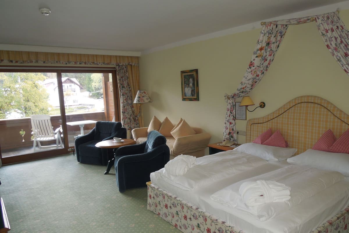 A hotel room with a bed, two armchairs, a sofa, a small table, and a balcony with a view of buildings and trees.