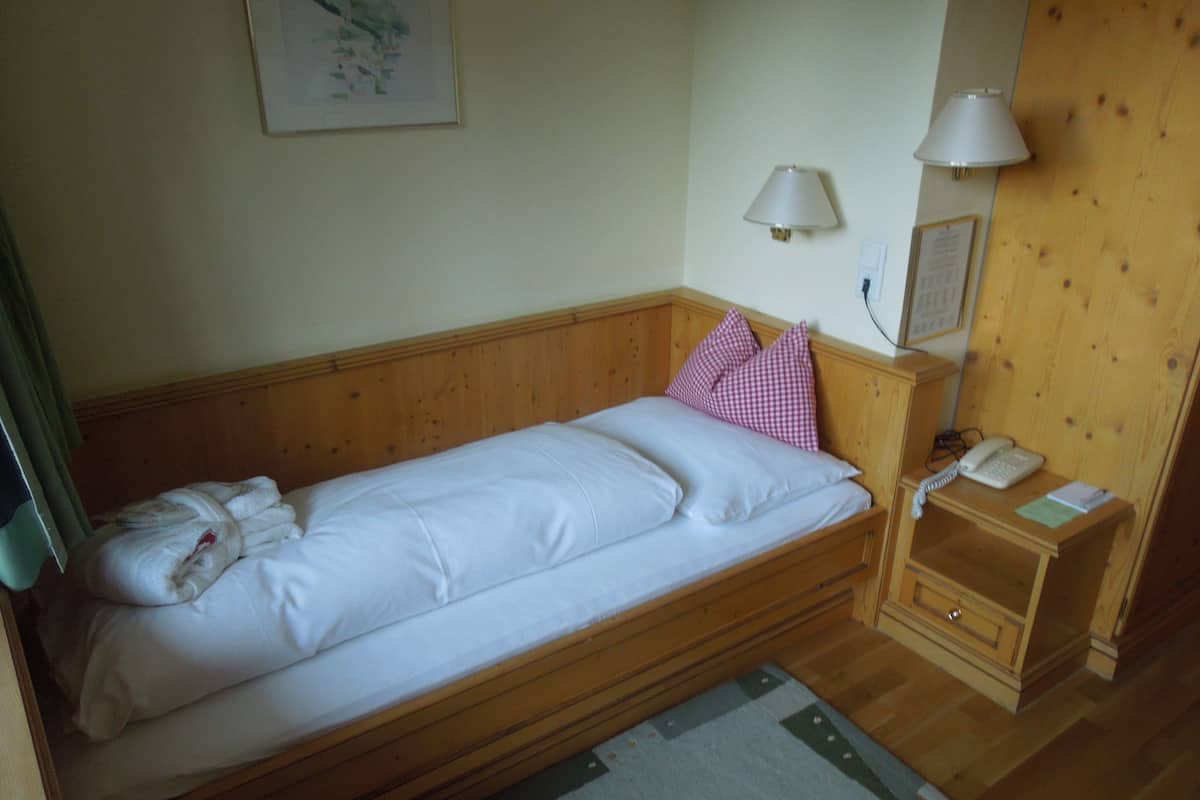 A single bed with white linens, a wooden headboard, a bedside table with a lamp, a phone, and a small cabinet.