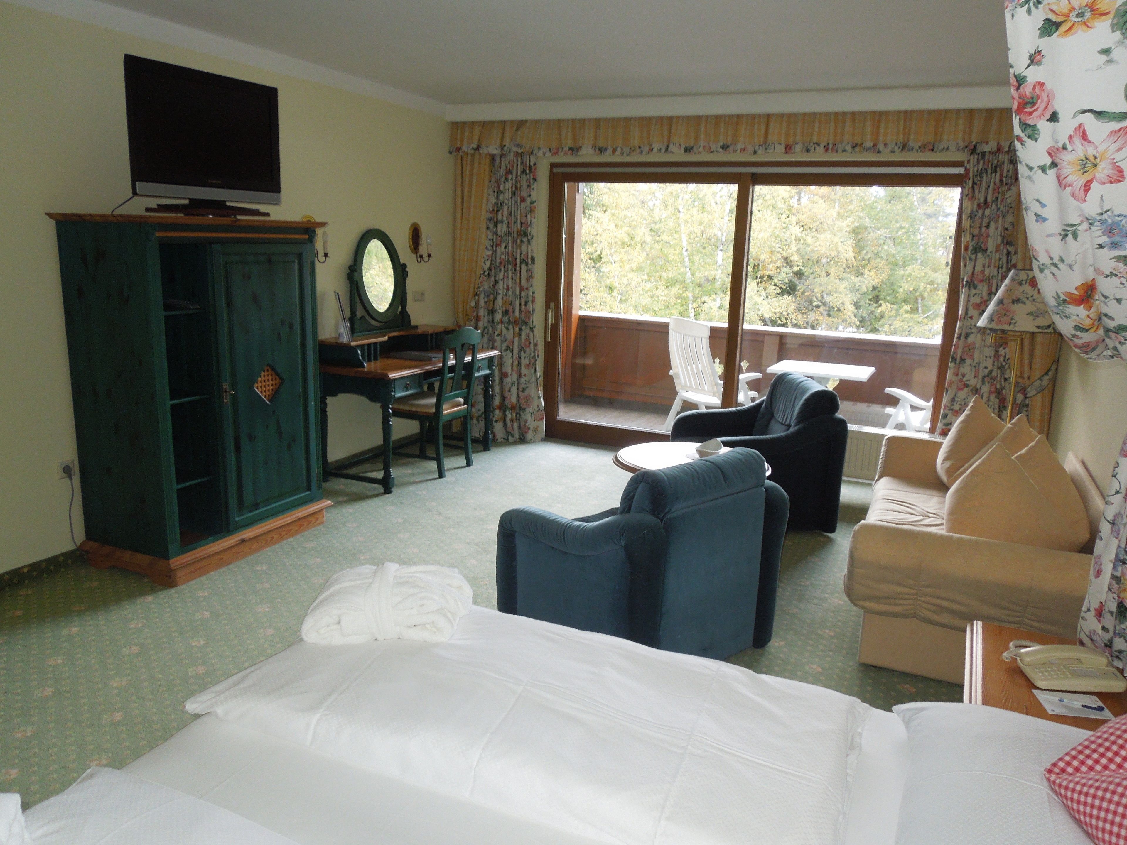 Ein Hotelzimmer mit Bett, Fernseher, Schreibtisch mit Stuhl, einer Sitzecke und einem Balkon mit Gartenmöbeln.