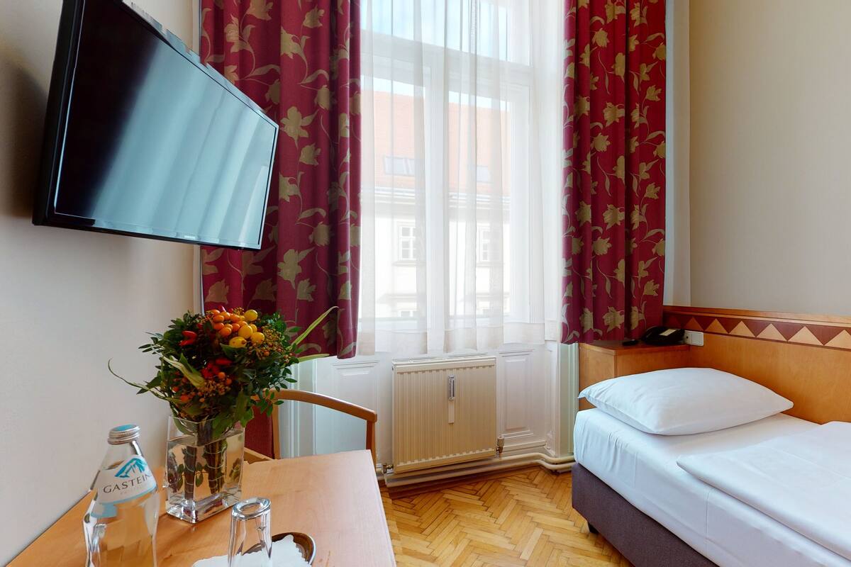 Ein Hotelzimmer mit Bett, Schreibtisch mit Blumenvase, Wasserflasche und flachbildschirm.
