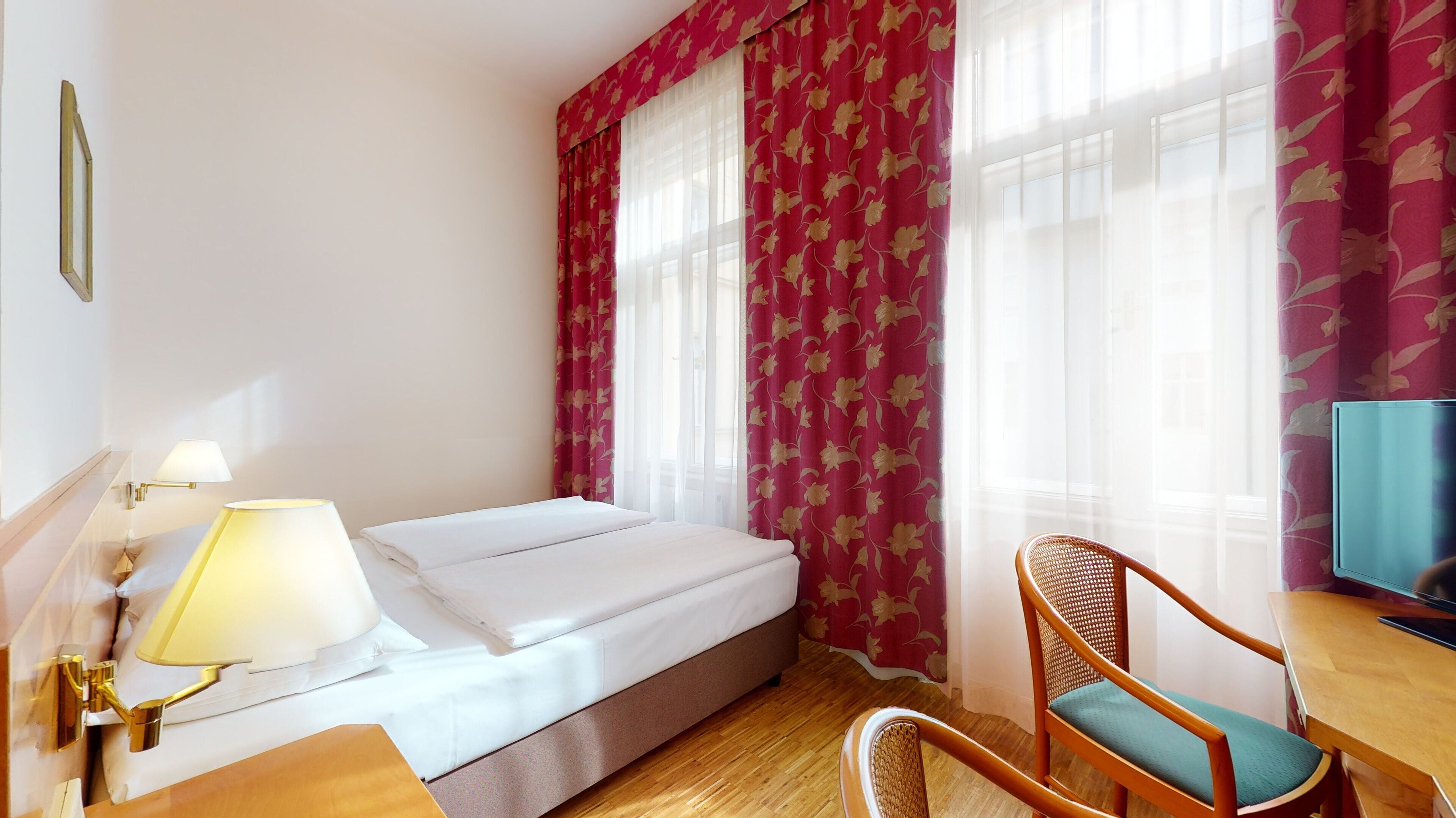 A hotel room with a bed, a desk with a chair, a television, and a window with curtains.