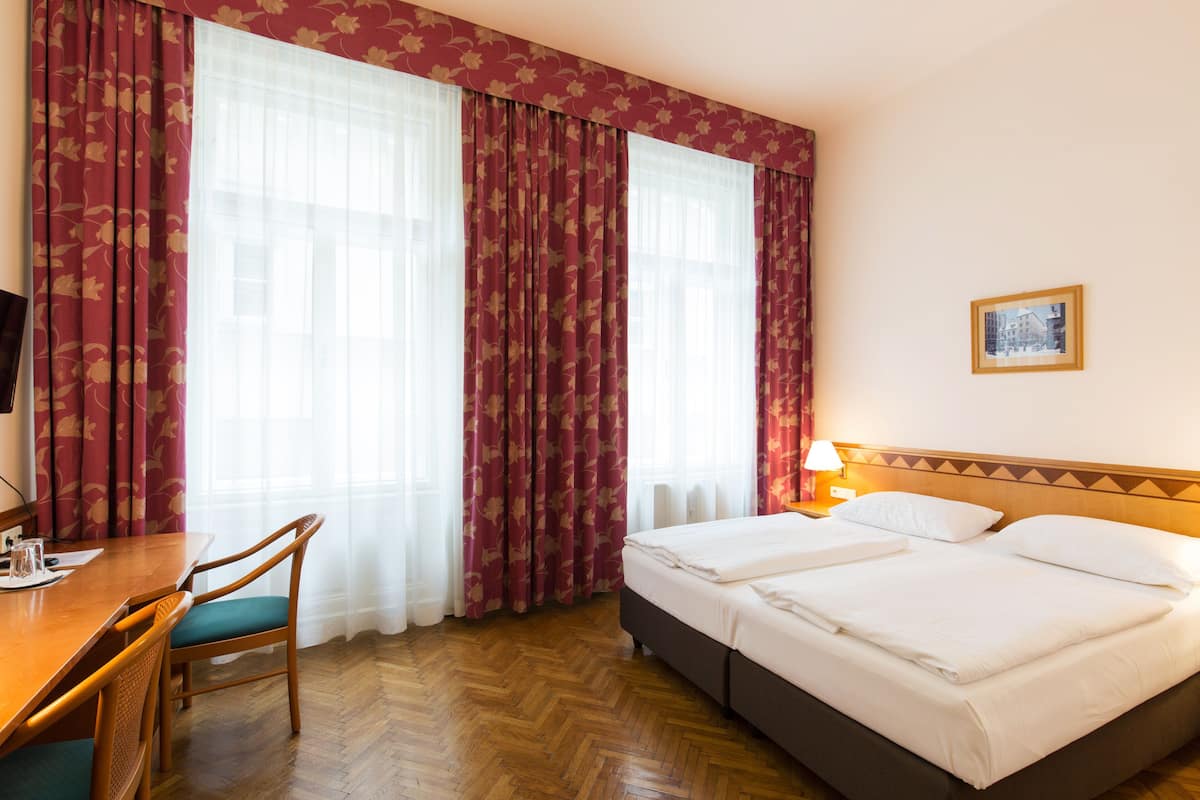 Ein Hotelzimmer mit zwei Betten, einem Holztisch mit Stühlen und großen Fenstern mit roten Vorhängen.
