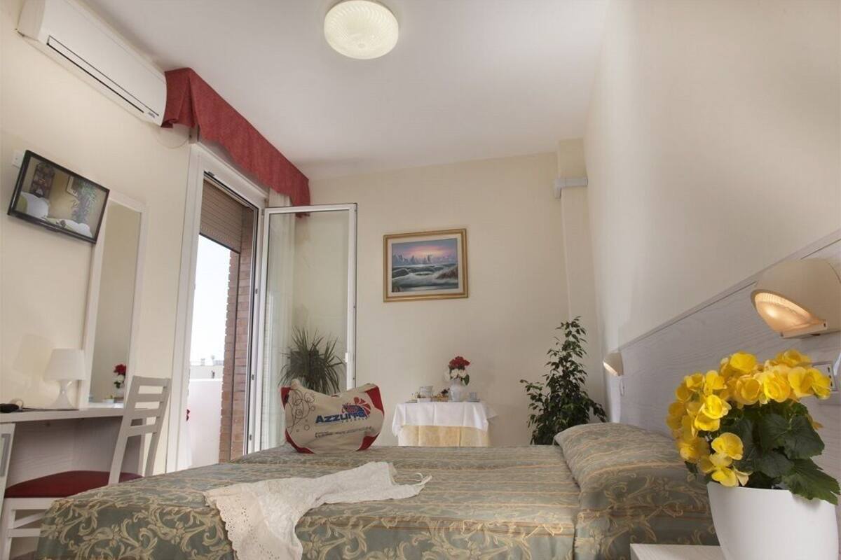 A hotel room with a bed, a desk, a chair, a table with a vase of flowers, and a painting on the wall.