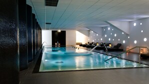A modern indoor swimming pool with a tiled floor, a glass barrier, and multiple lounge chairs along the side. - Open Village Sports Hotel & SPA Club (Guimarães)