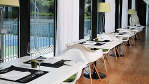 A modern dining area with long tables, white chairs, and large windows offering a view of a tennis court. - Open Village Sports Hotel & SPA Club (Guimarães)