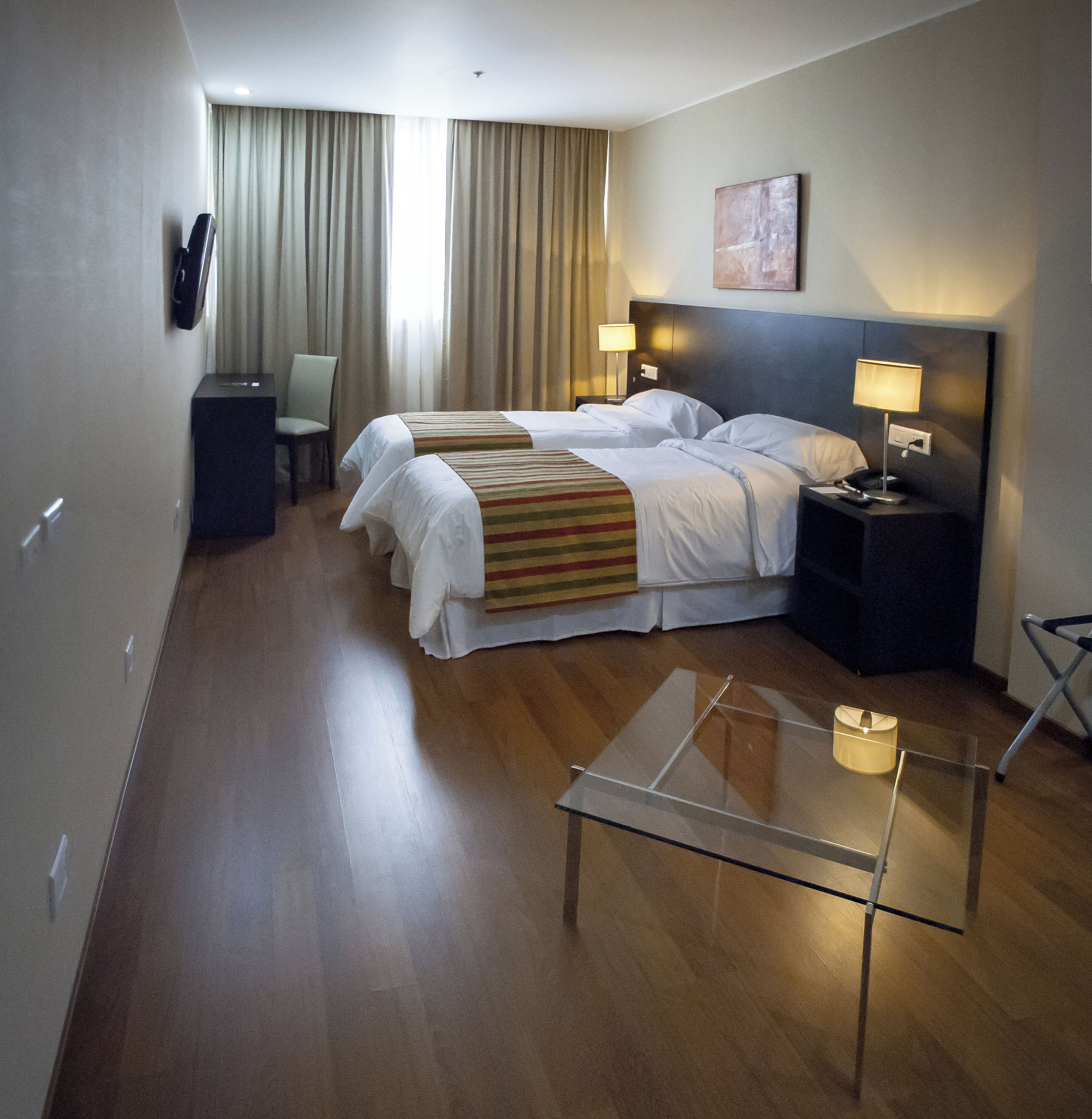 A hotel room with two beds, a desk, a chair, a lamp, and a glass table.