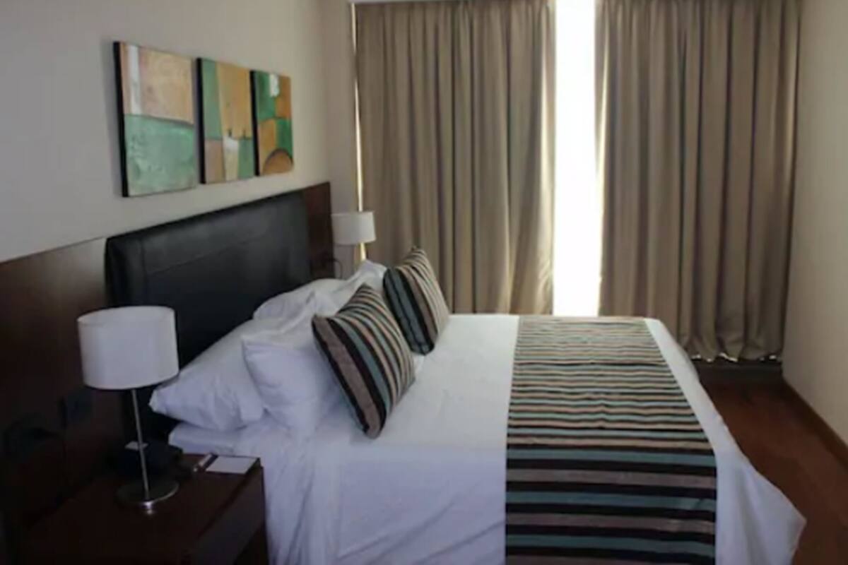 A hotel room with a large bed, a nightstand with a lamp, and a view of the outside through curtains.