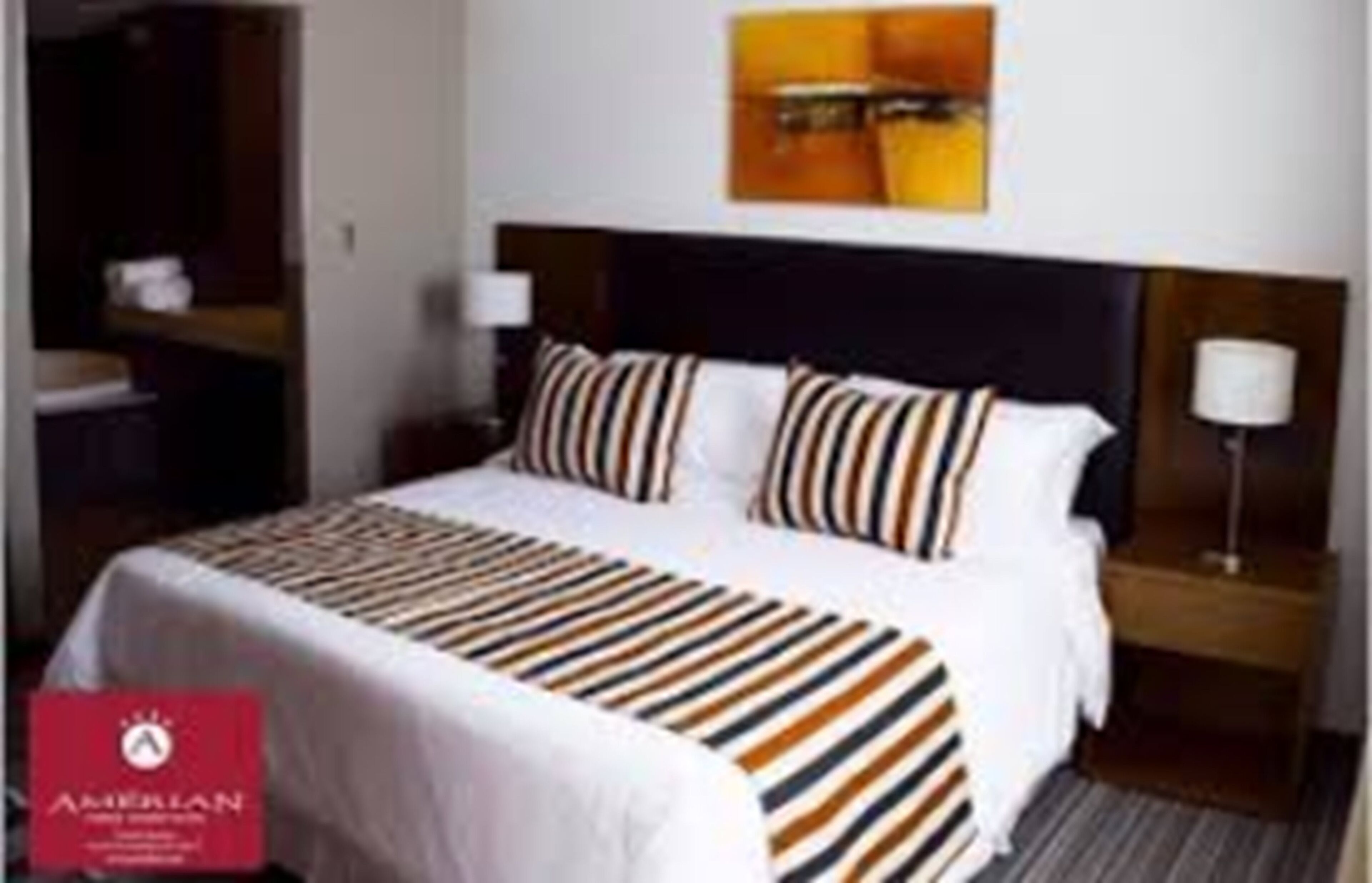 A hotel room with a neatly made bed, a headboard, a bedside table with a lamp, and a wall-mounted artwork.