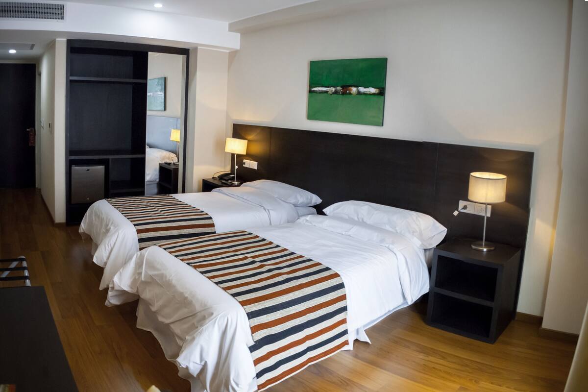 A hotel room with two beds, a wooden floor, a green abstract painting, and a nightstand with a lamp.