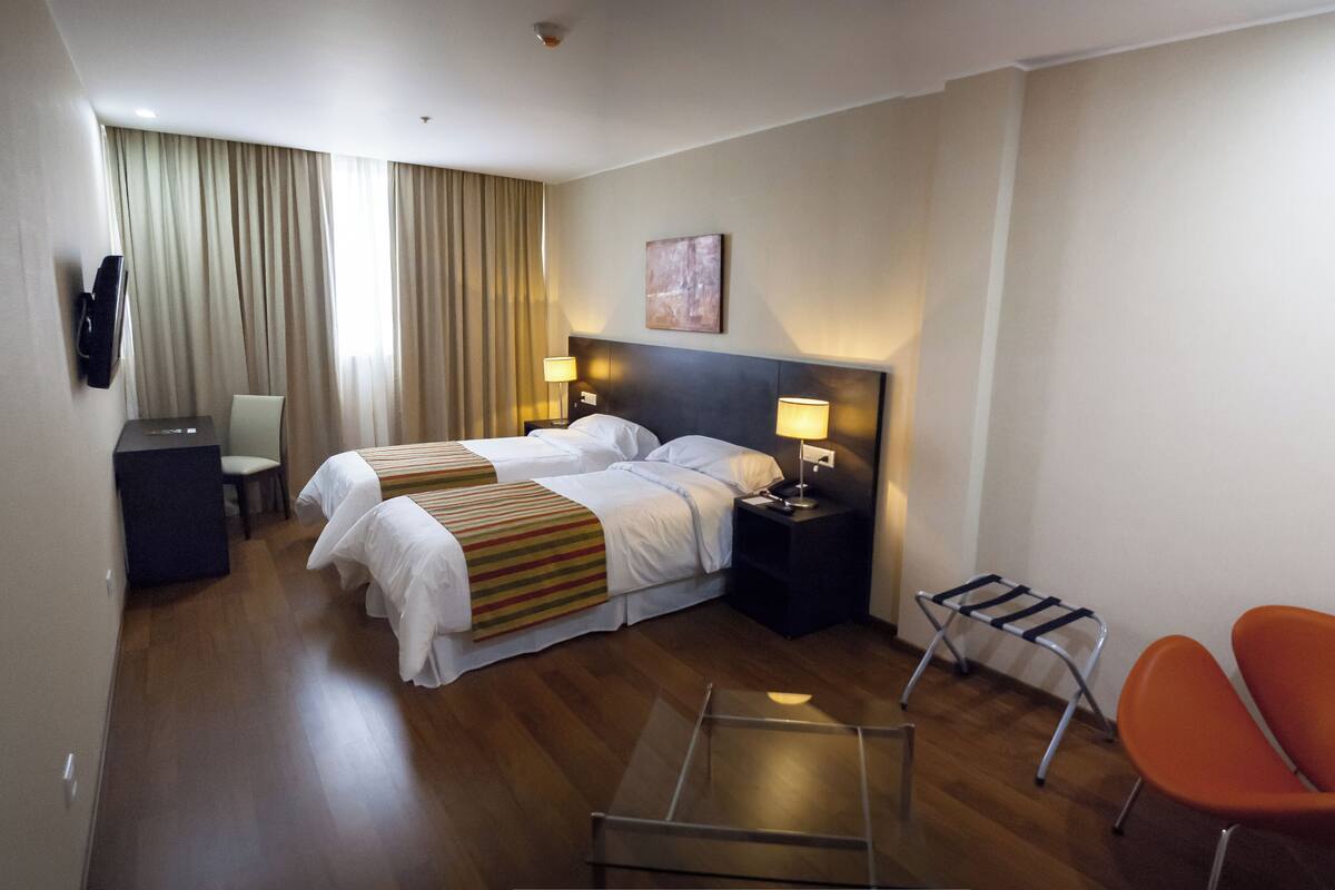 A hotel room with two beds, a desk, a chair, a lamp, and a small table.