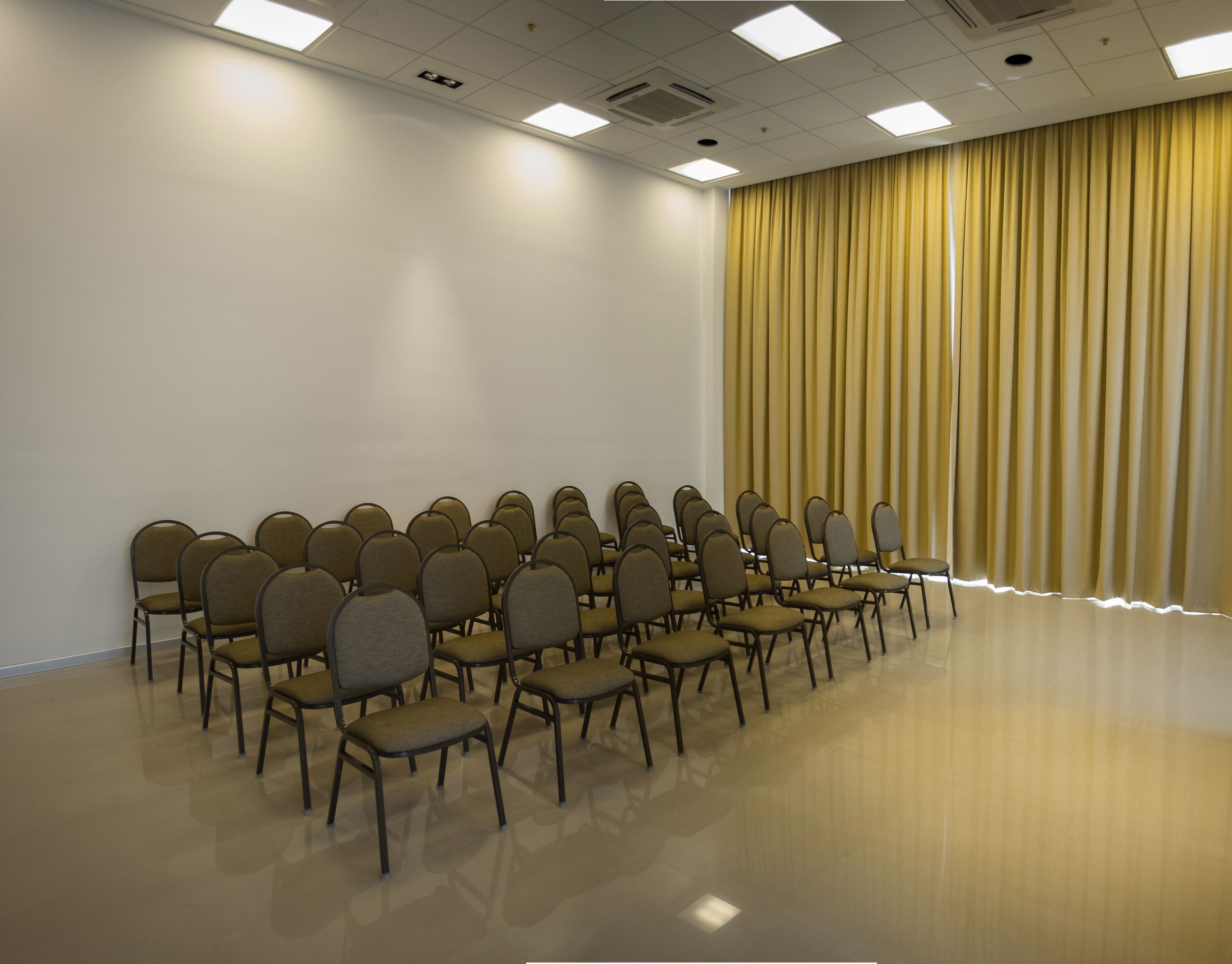 Un salón de conferencias con filas de sillas, una pared blanca lisa y una cortina amarilla.