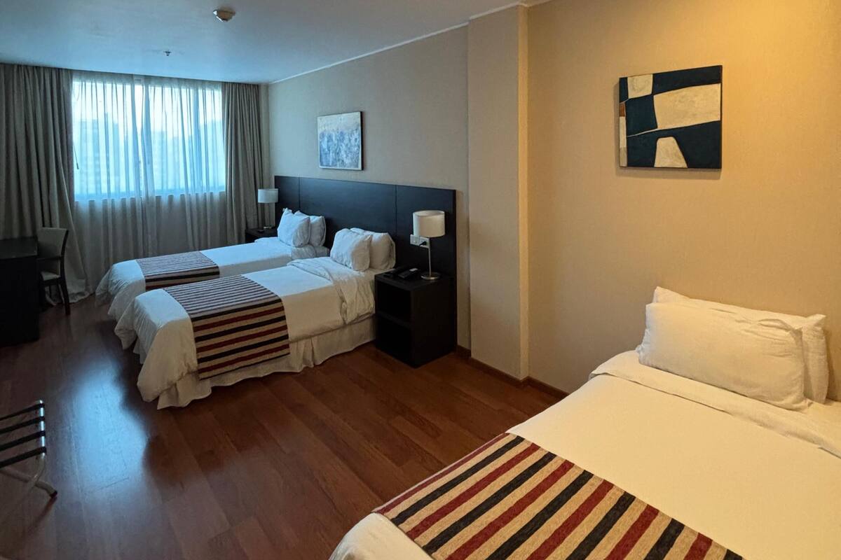 A hotel room with two beds, a desk, a chair, a nightstand with a lamp, and a painting on the wall.