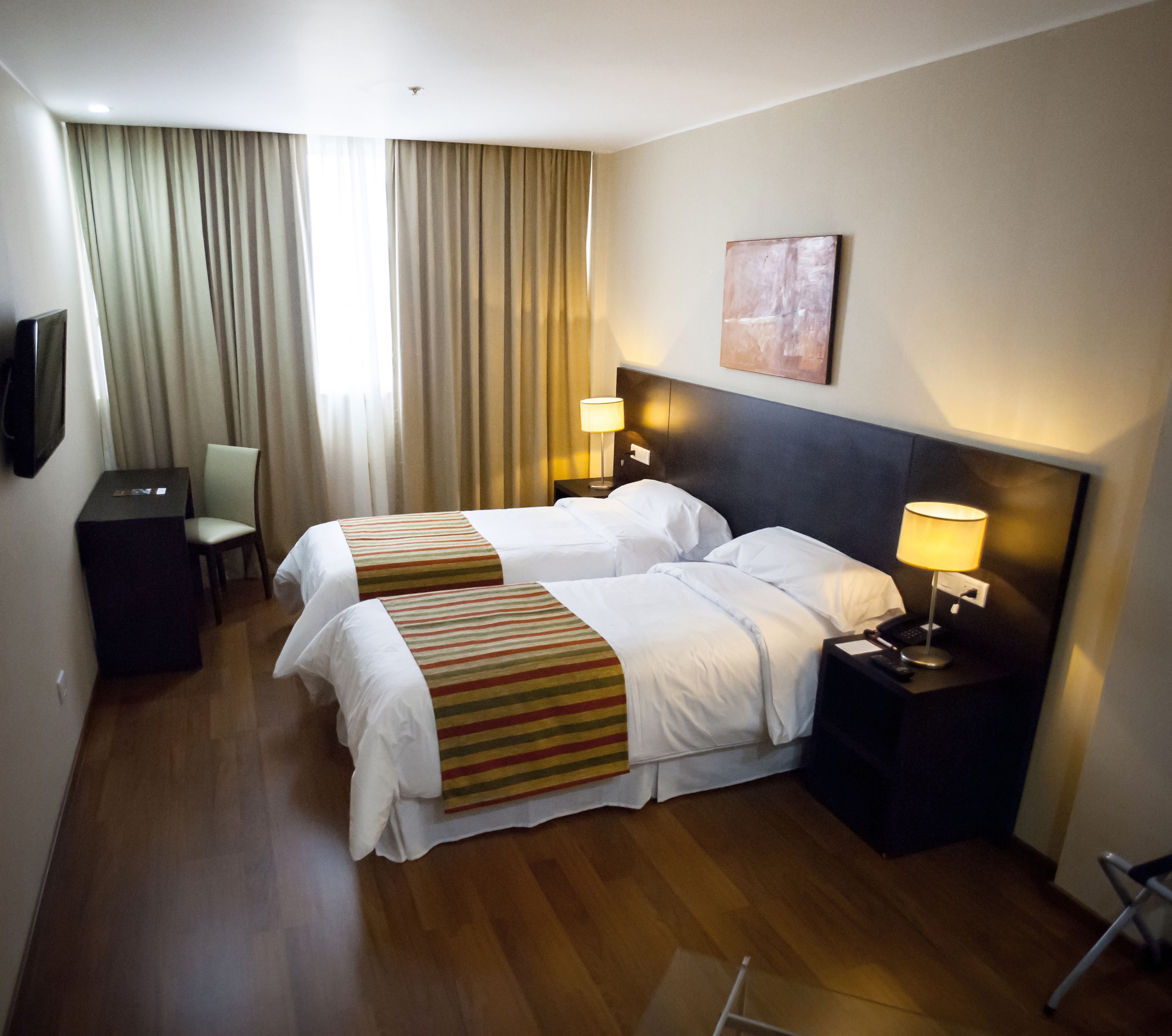 A hotel room with two beds, a desk, a chair, a TV, and a painting on the wall.