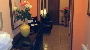 Um corredor de hotel com um vaso amarelo de flores, uma lixeira e uma mesinha com um abajur.