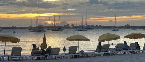 A beach with lounge chairs and umbrellas, boats in the background, and a sunset.