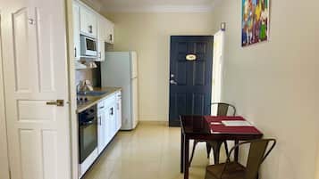 A kitchen with white cabinets, a microwave, and a refrigerator. There is a dining table with chairs and a door with a number sign.