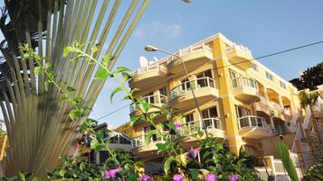 A multi-story yellow building with balconies, surrounded by greenery and flowers.