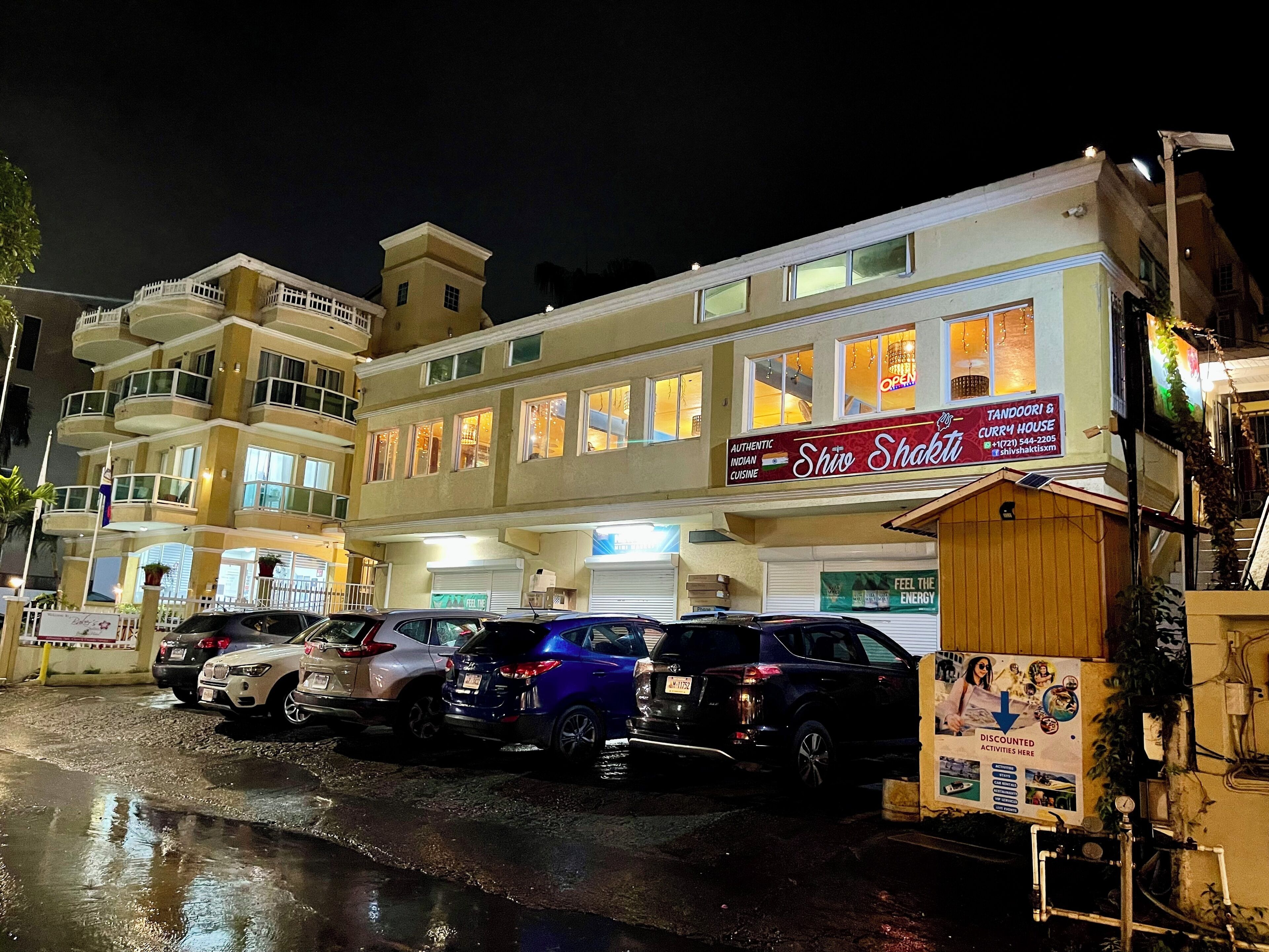 Een meerlaags gebouw met een restaurant genaamd “Shiv Shakti” op de begane grond. Er staan meerdere auto’s voor geparkeerd en het gebouw heeft meerdere balkons.