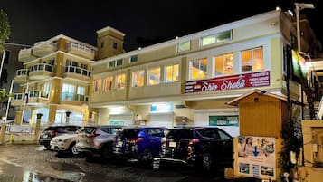 Een meerlaags gebouw met een restaurant genaamd “Shiv Shakti” op de begane grond. Er staan meerdere auto’s voor geparkeerd en het gebouw heeft meerdere balkons.