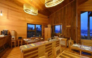A wooden cabin interior with a dining area, kitchen, and large windows offering a view of the city.