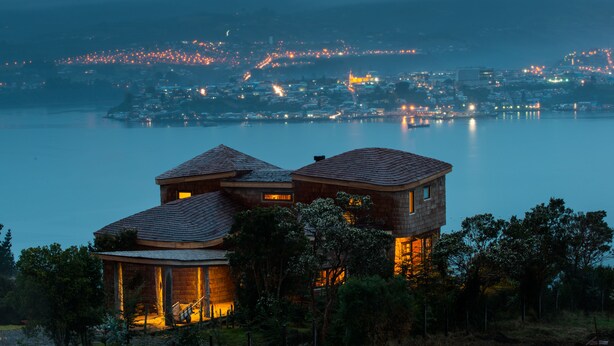 A two-story wooden house with a balcony, surrounded by trees, overlooking a calm lake with a cityscape in the background. - OCIO Territorial Hotel (Castro)