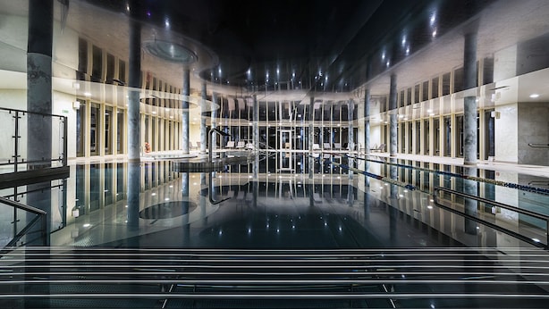 A spacious indoor swimming pool with clear glass walls, a central water feature, and a modern architectural design. - Hotel Kiston (Suleczyno)