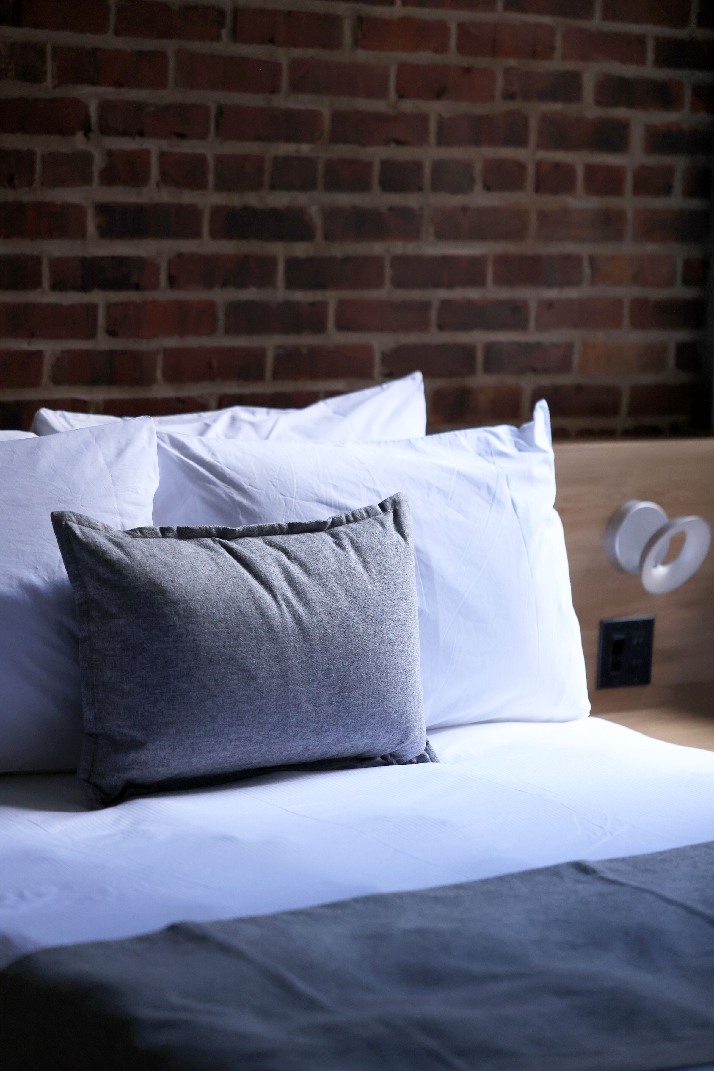 Uma cama com almofadas brancas e cinzentas, uma cabeceira de madeira e uma parede de tijolos ao fundo.