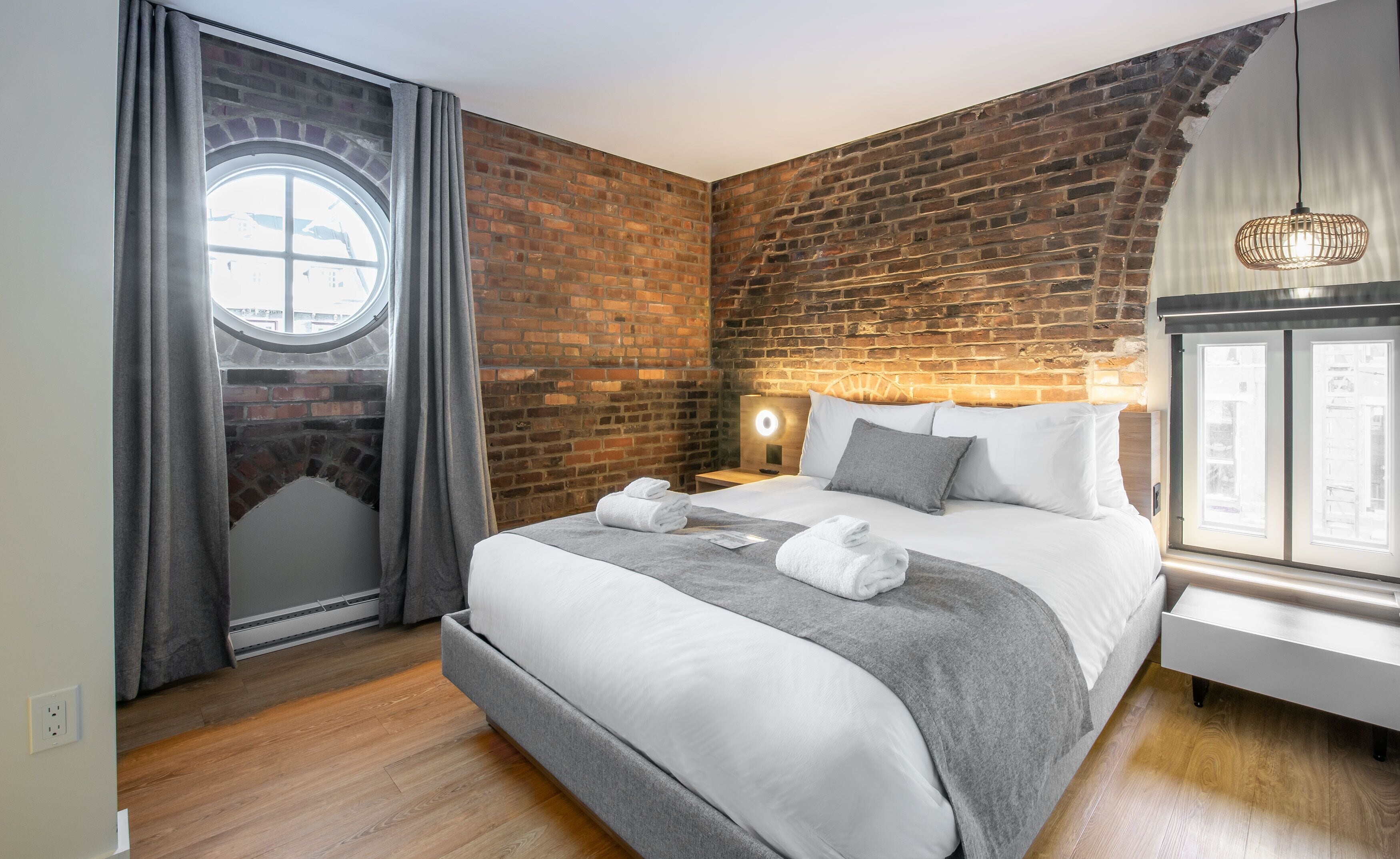 Ein Hotelzimmer mit Backsteinwand, einem Rundfenster, einem Bett mit weißer Bettwäsche, einem Nachttisch mit Lampe und Blick ins Freie.