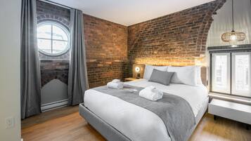 Ein Hotelzimmer mit Backsteinwand, einem Rundfenster, einem Bett mit weißer Bettwäsche, einem Nachttisch mit Lampe und Blick ins Freie.