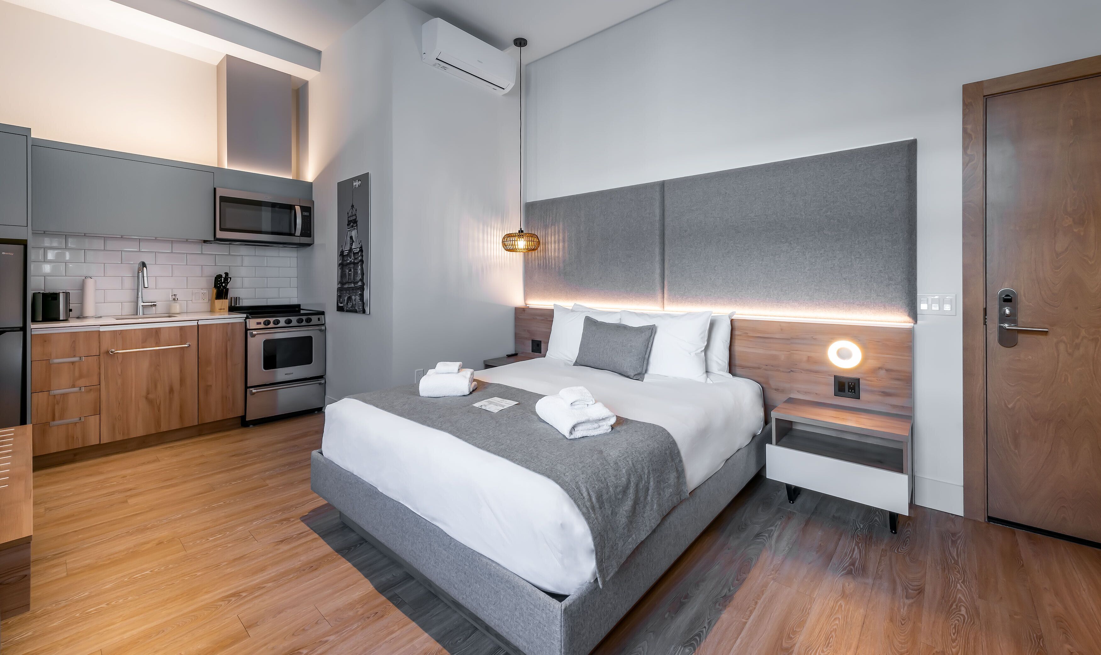 Une chambre d’hôtel moderne avec un coin cuisine, un lit recouvert de draps blancs et une porte en bois.
