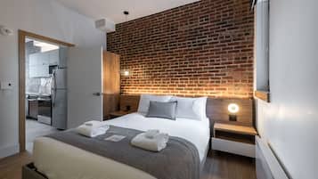 A modern hotel room with a brick accent wall, a neatly made bed, a small kitchen area, and a view of the adjacent room.