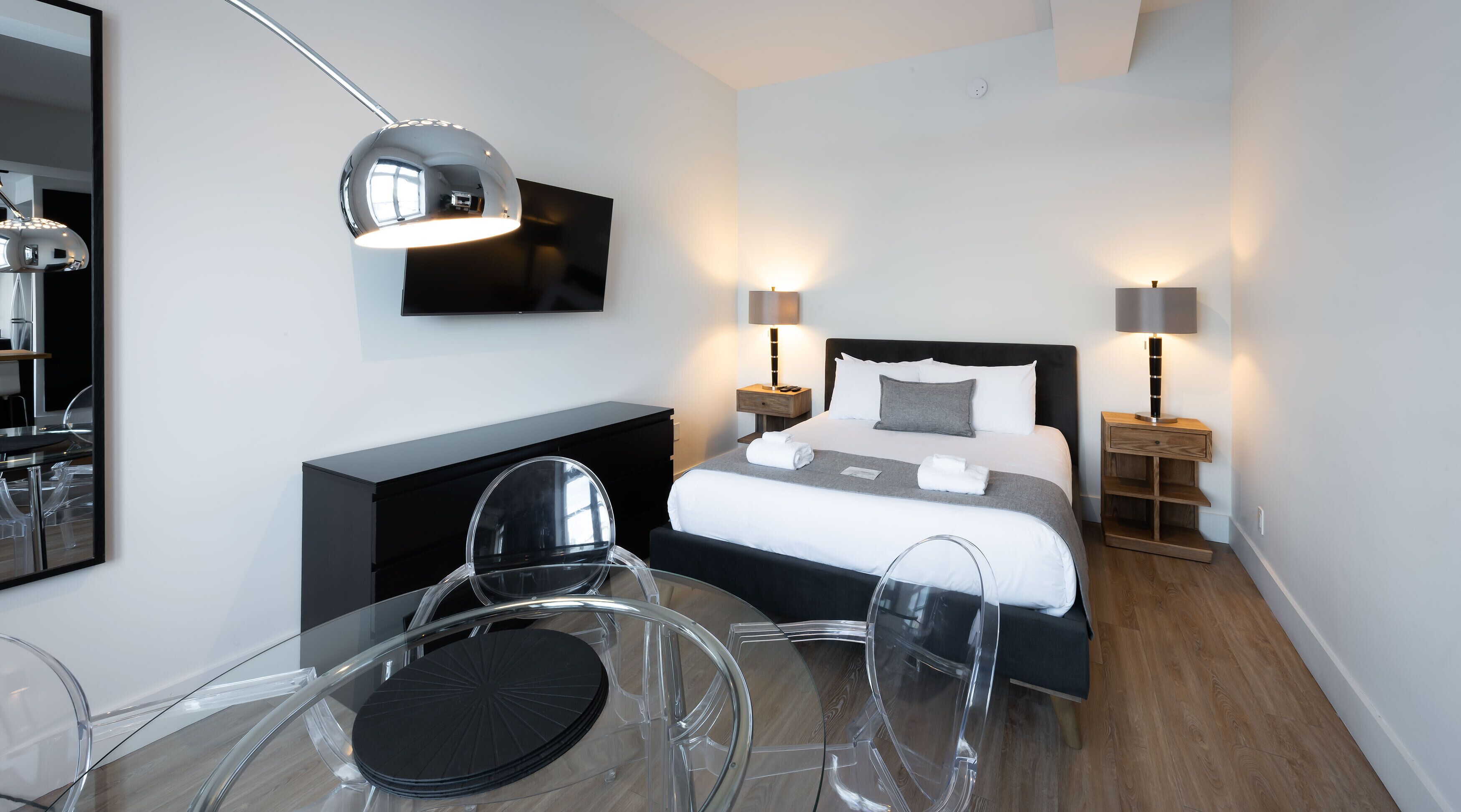 A modern hotel room with a bed, bedside lamps, a TV, a desk with a chair, and a mirror.