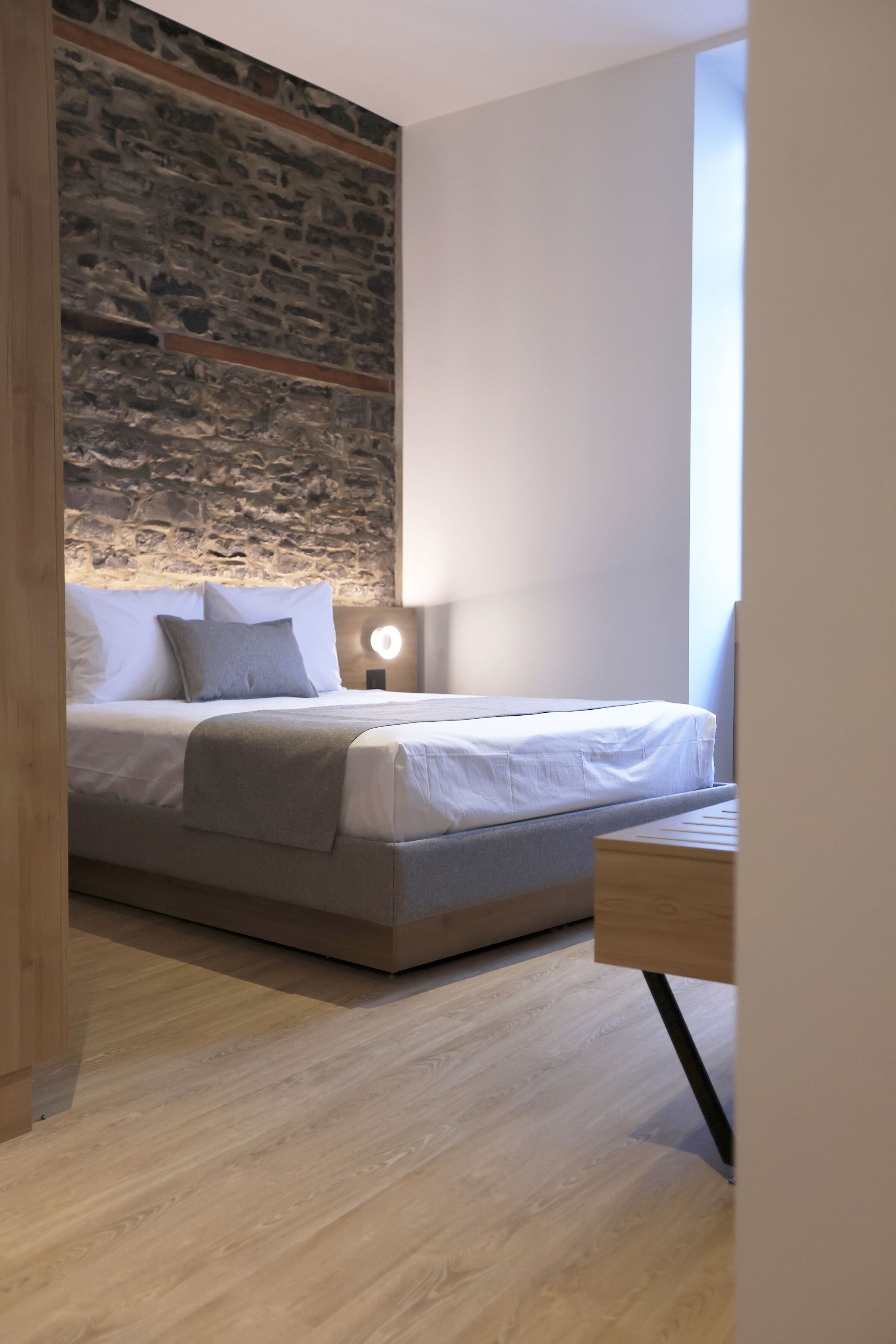 Une chambre moderne avec un mur d’accent en pierre, un lit avec une literie blanche, une table de chevet avec une lampe et une vue sur l’extérieur.