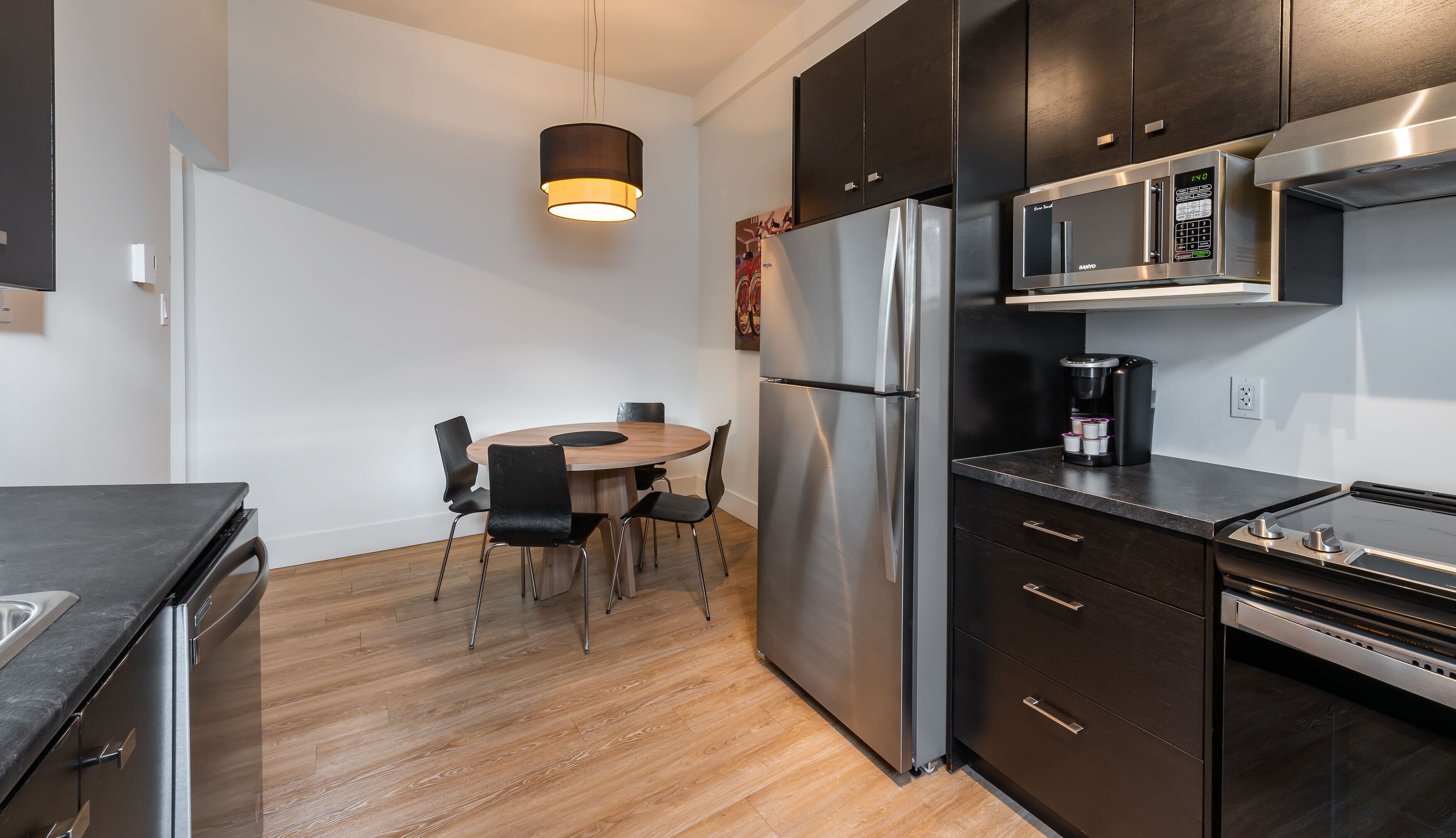 A modern kitchen with a stainless steel refrigerator, a coffee maker, and a microwave. There is a wooden dining table with chairs.