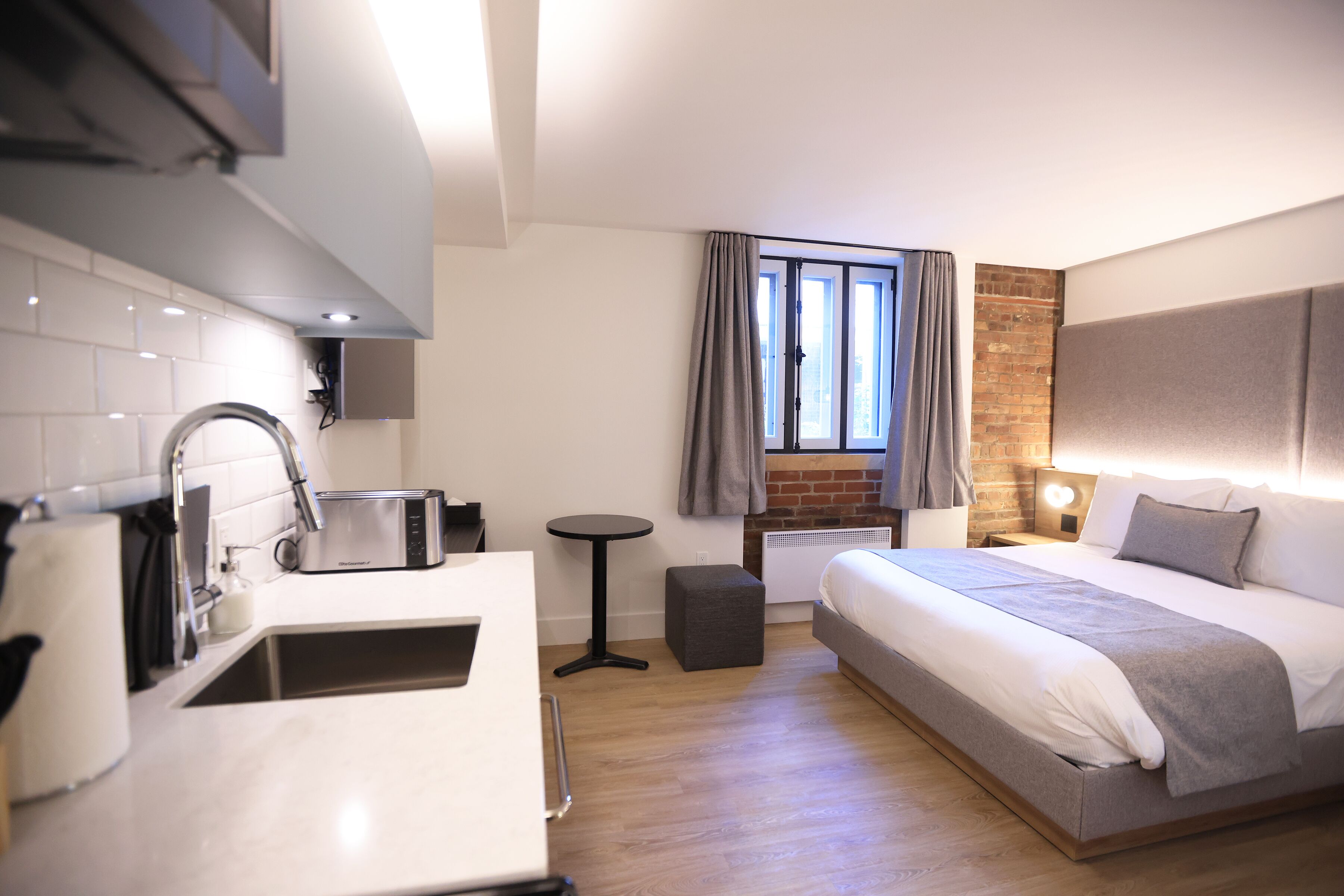 Habitació d'hotel moderna amb petita cuina, un llit amb capçalera gris, una taula rodona i una finestra amb cortines.