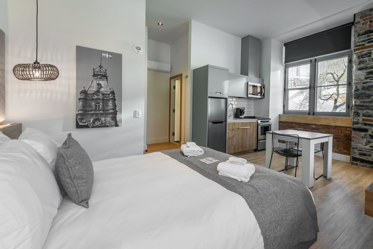 A modern hotel room with a bed, a desk, a kitchenette, and a large window with a view.