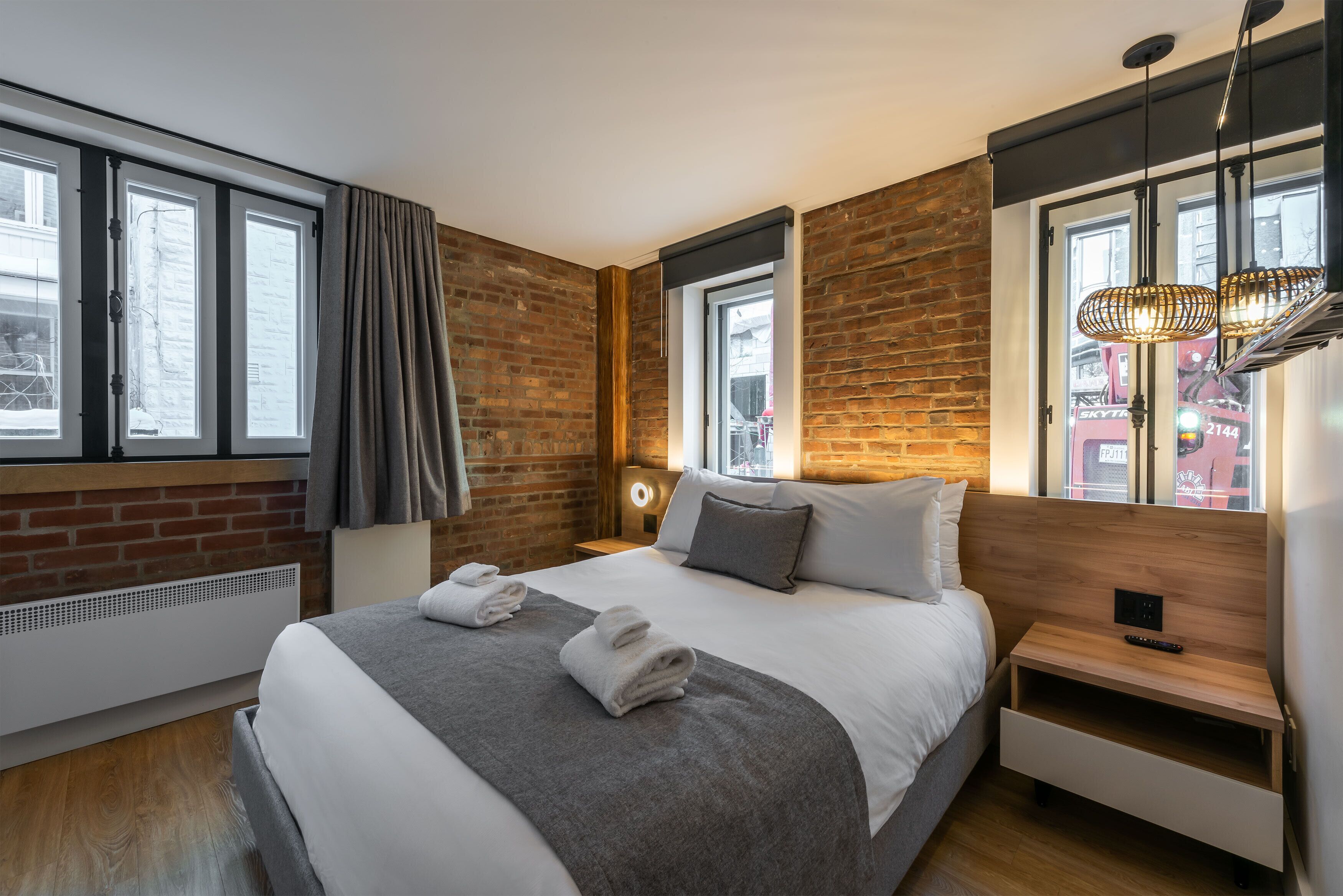 A modern hotel room with a large bed, a wooden headboard, a nightstand, and a window with a view of the city.
