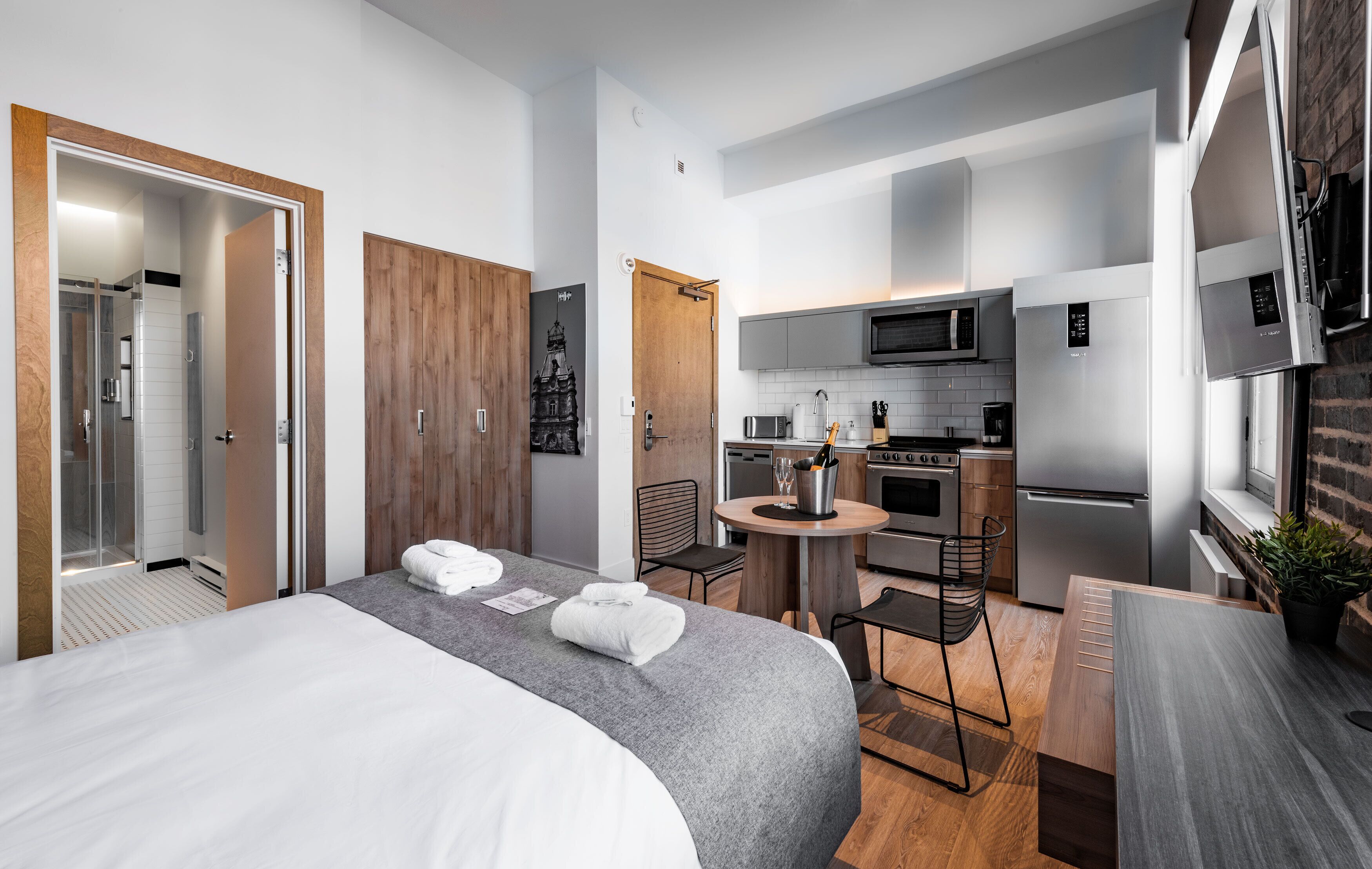 A compact hotel room with a bed, a small kitchenette, a dining area, and a bathroom with a shower.