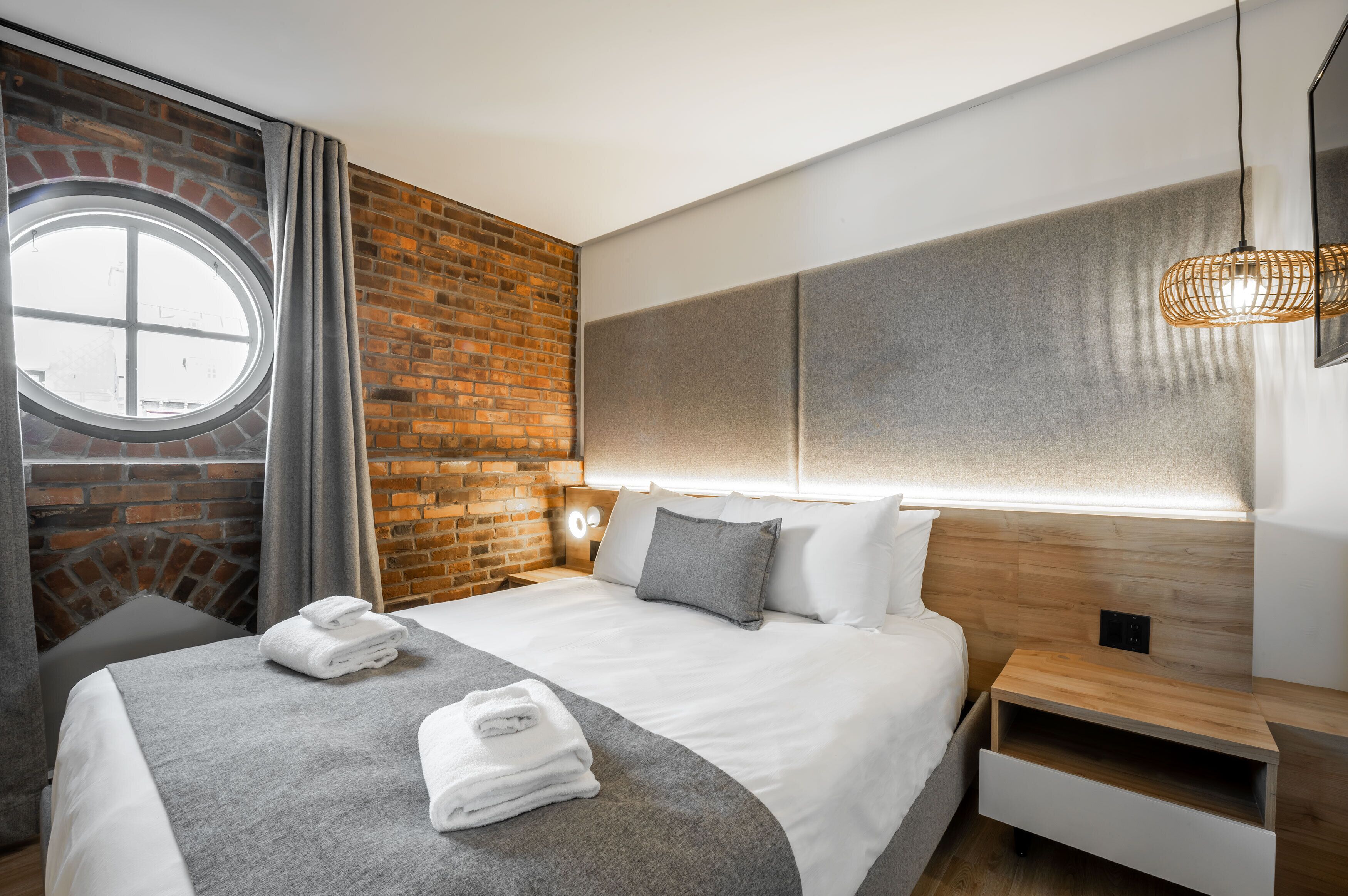 A modern hotel room with a brick accent wall, a round window, a neatly made bed with white linens, and a bedside table with a lamp.