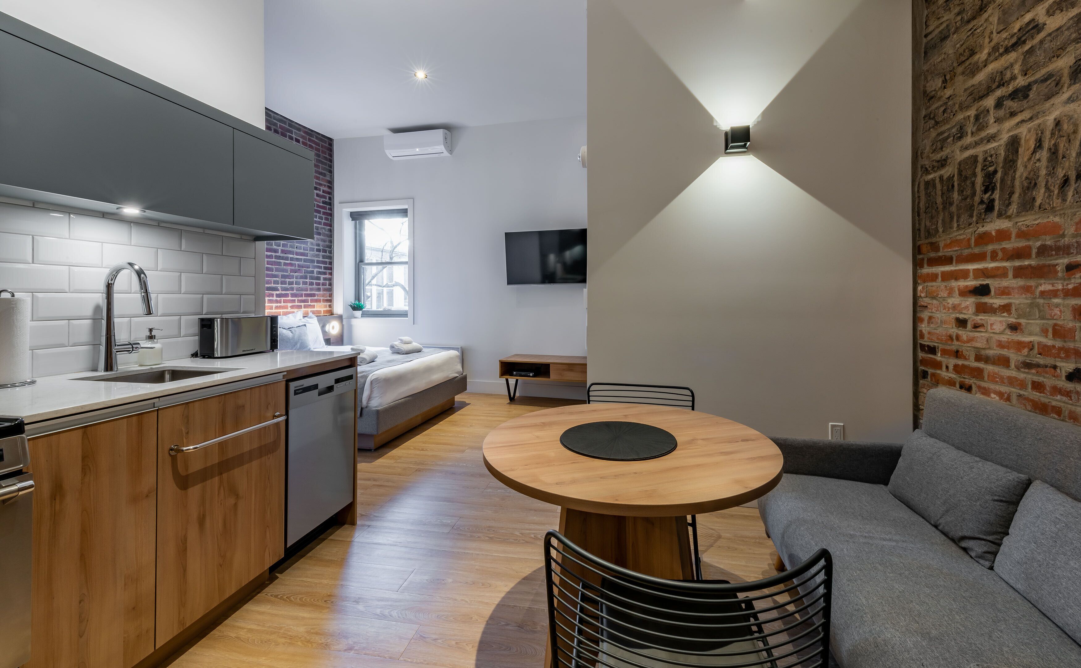 Una cocina moderna con lavavajillas incorporado, una zona de comedor con una mesa redonda de madera y sillas de metal, y un sofá gris.