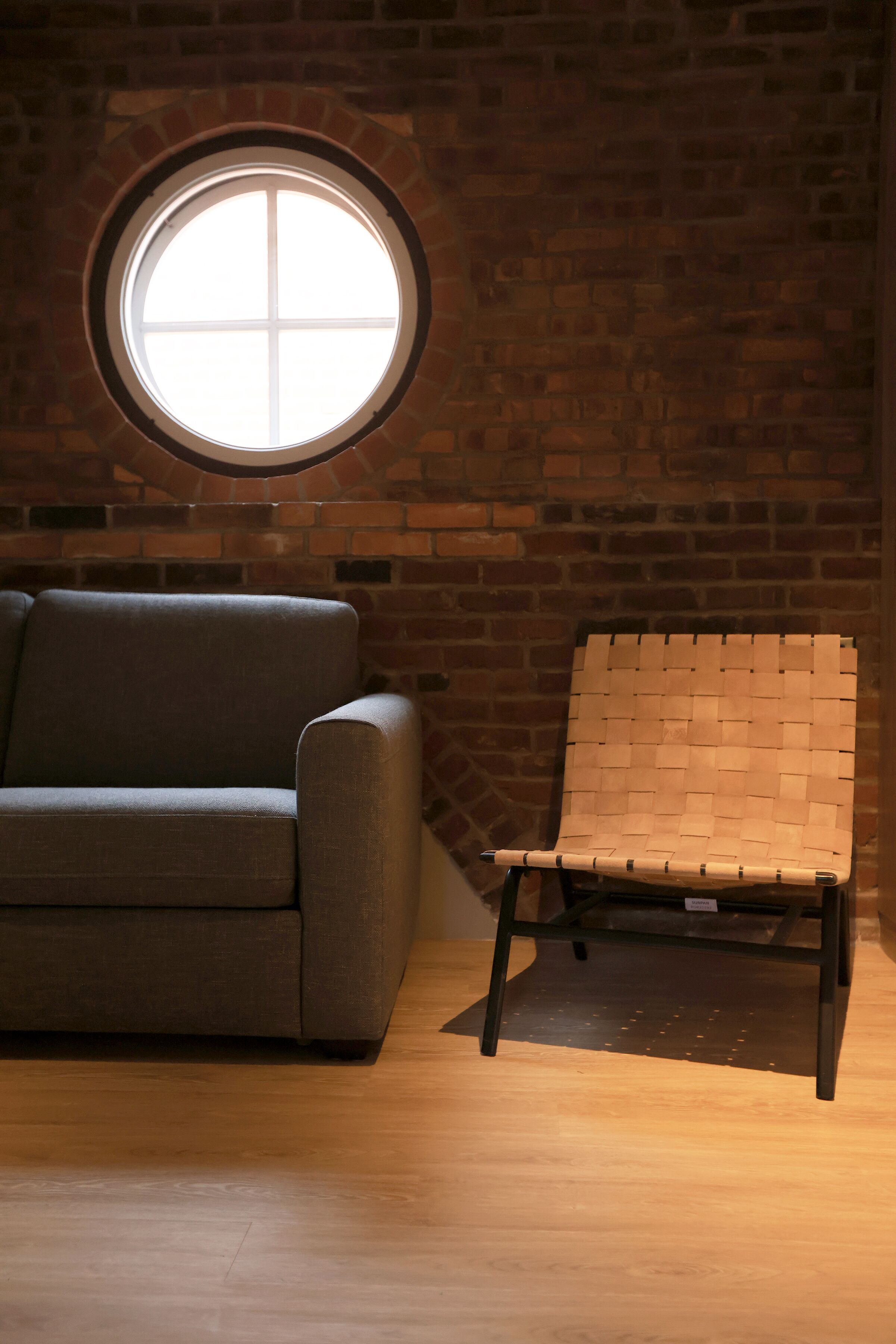 A room with a brick wall, a round window, a grey sofa, and a woven chair.