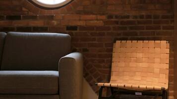 A room with a brick wall, a round window, a grey sofa, and a woven chair.