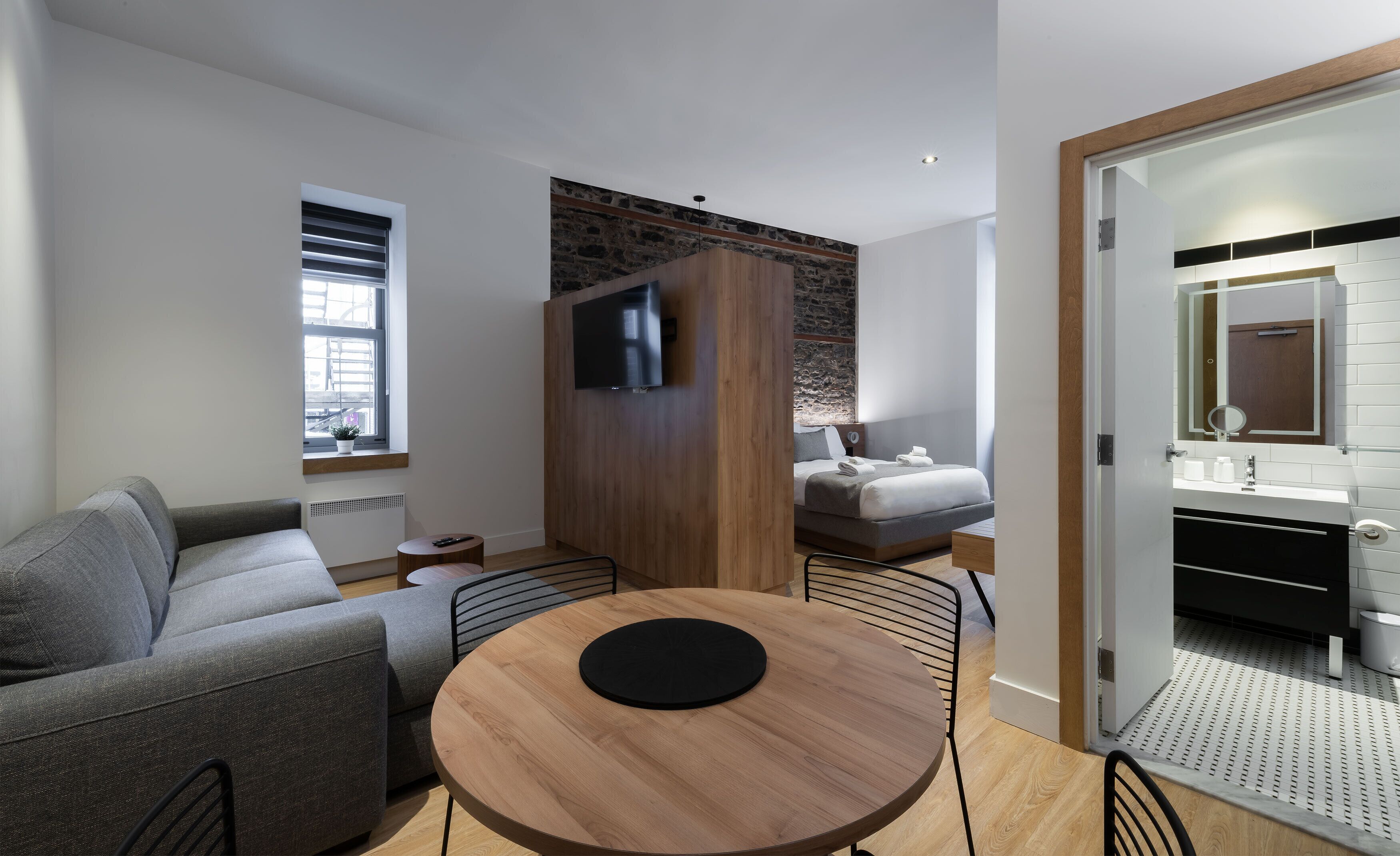 A modern hotel room with a grey sofa, a round wooden table, a TV mounted on a wooden panel, a bed with a grey headboard, and a bathroom with a mirror and sink.