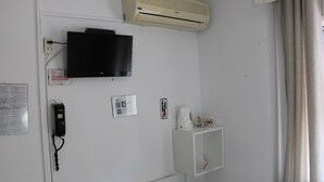 Ein Zimmer mit einem an der Wand befestigten Fernseher, einem weißen Regal mit Wasserkocher und Tassen, einem Telefon und einer Wandklimaanlage.