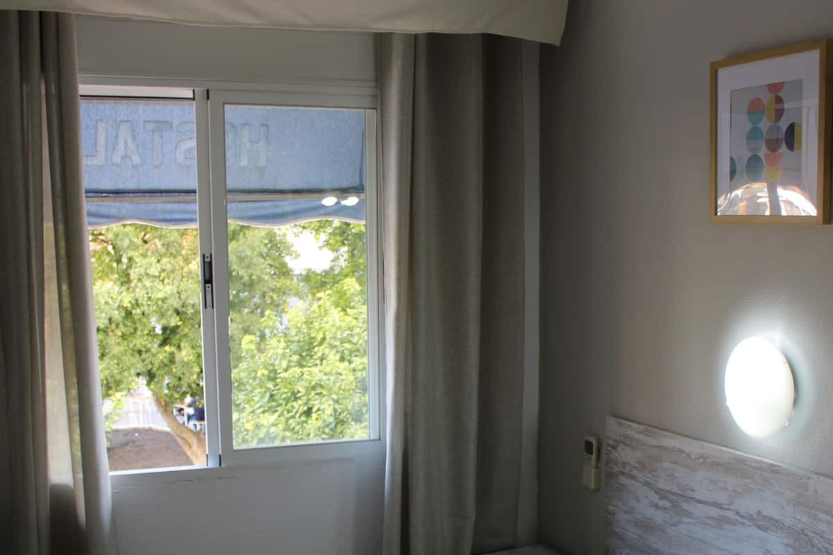 Um quarto com cama, mesa de cabeceira, janela com cortinas e luminária na parede.