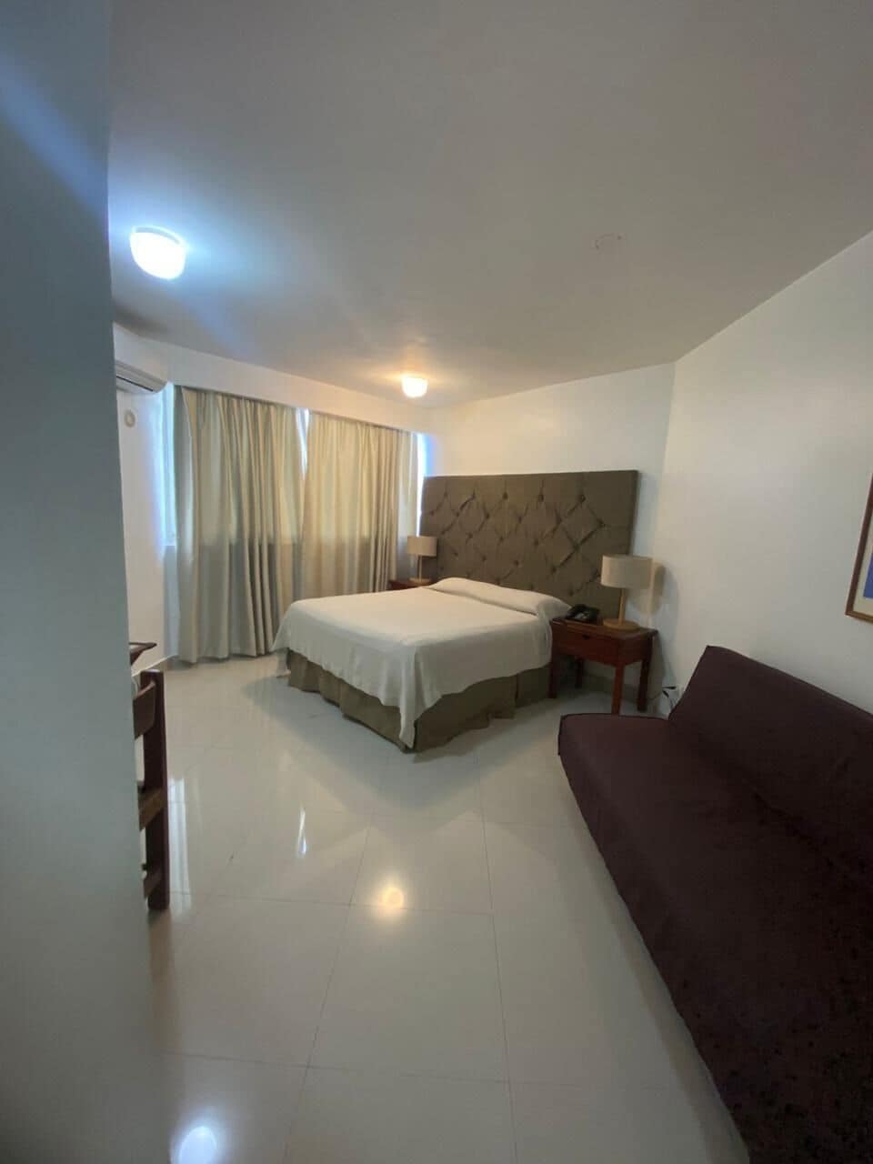 A hotel room with a bed, bedside tables, a sofa, a chair, and a window with curtains.
