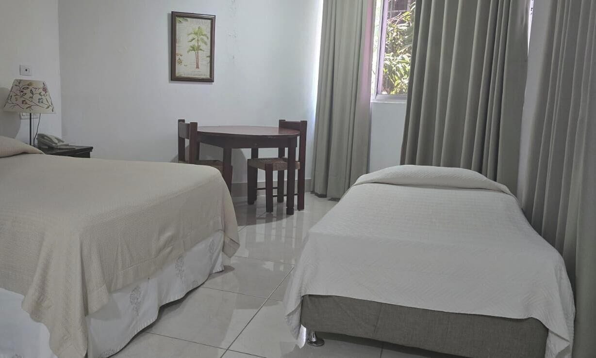 A hotel room with two beds, a small table, a chair, a lamp, and a window with curtains.