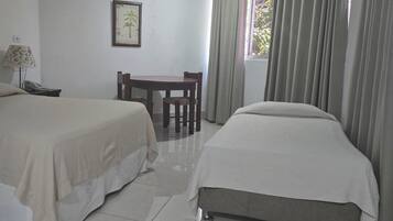 A hotel room with two beds, a small table, a chair, a lamp, and a window with curtains.