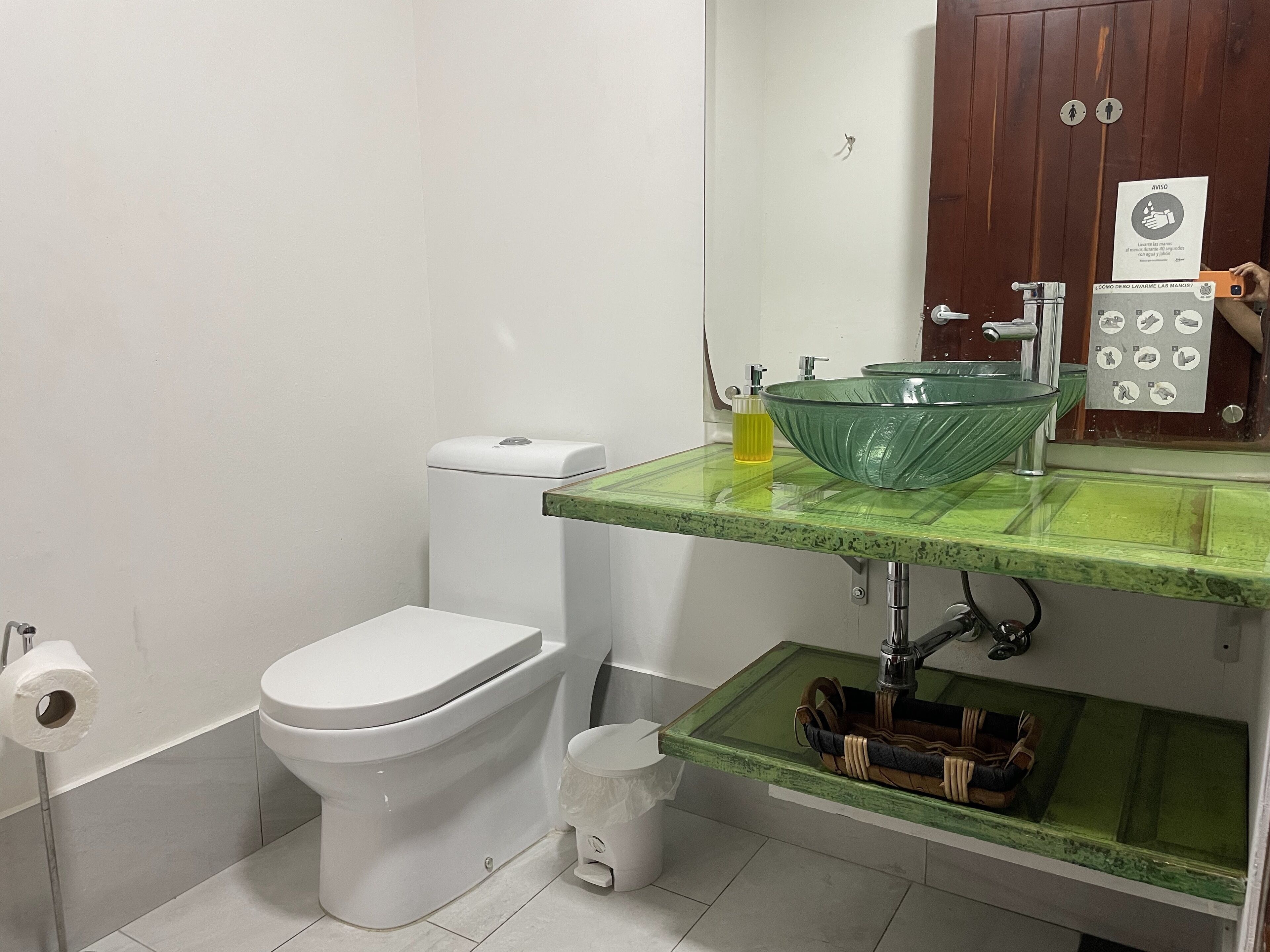 Une salle de bain avec des toilettes blanches, un lavabo vert doté d’un receveur en verre et un meuble en bois au-dessus du lavabo.