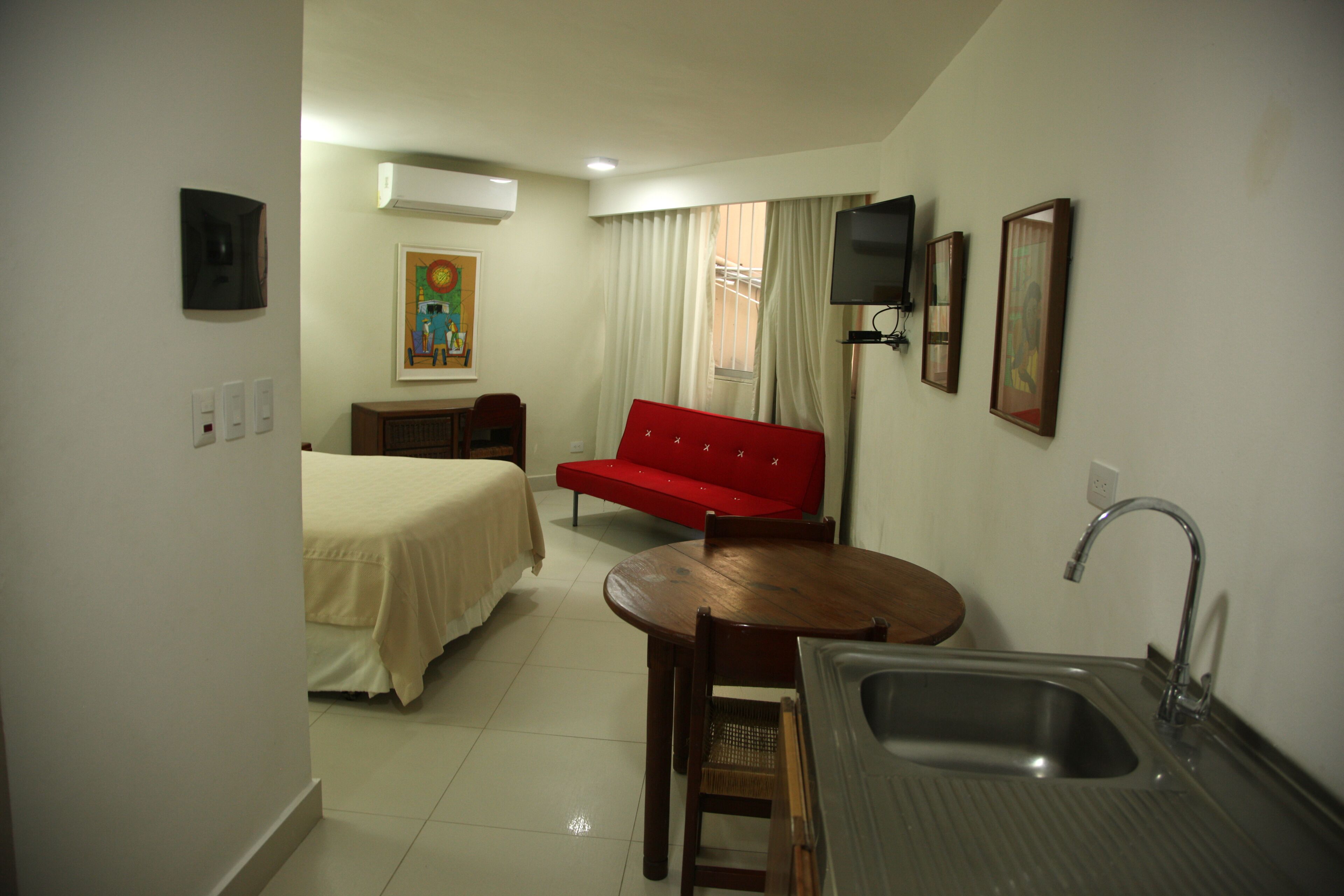 A compact hotel room with a bed, a red sofa, a small dining table, a sink, and a TV mounted on the wall.